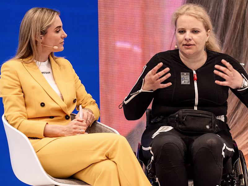 TV-Moderatorin Anna Kraft und eine Exopulse Mollii Suit Anwenderin im Rollstuhl auf der Show-Bühne von Ottobock auf der OTWorld 2024 in Leipzig.