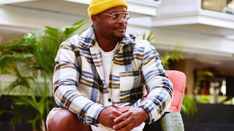 A young man with a prosthetic leg sitting in front of a building, wearing glasses, a yellow cap, and a blue and brown checked shirt.