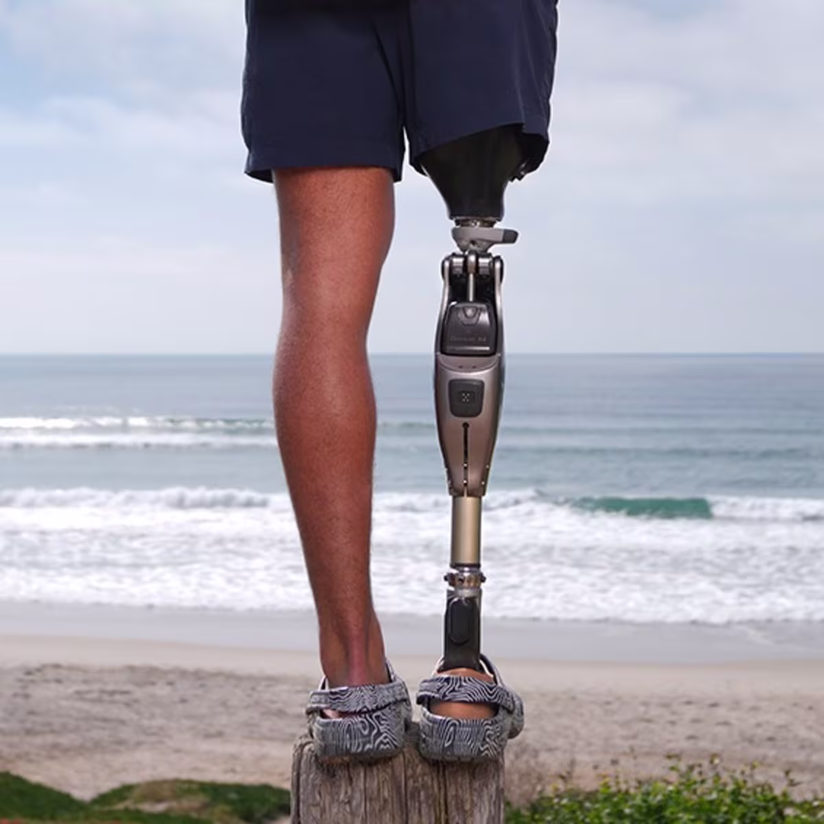 Das Bild zeigt den Unterkörper einer amputierten Person. Am rechten Bein trägt die Person das mechatronische Prothesenkniegelenk Genium X4 von Ottobock. Im Hintergrund sind Strand und Meer zu sehen.