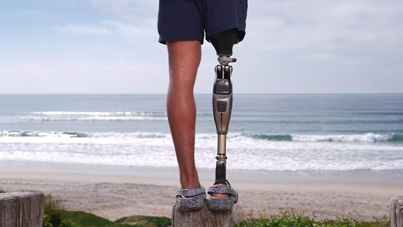 Das Bild zeigt den Unterkörper einer amputierten Person. Am rechten Bein trägt die Person das mechatronische Prothesenkniegelenk Genium X4 von Ottobock. Im Hintergrund sind Strand und Meer zu sehen.