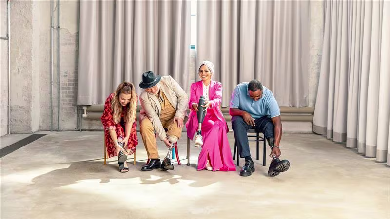 A person with a prosthetic leg stands on a wooden post overlooking a sandy beach and ocean waves, wearing shorts and casual shoes.
