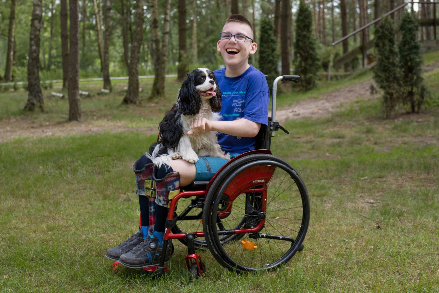 Nastolatek z MPD siedzi na wózku inwalidzkim. Na kolanach siedzi mu pies.