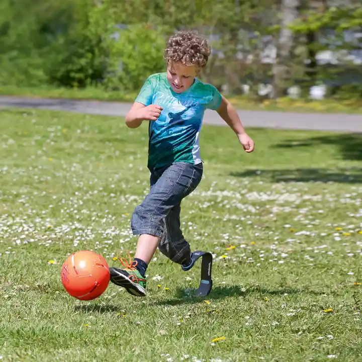 Kind mit Sportprothese spielt Fußball im Garten.