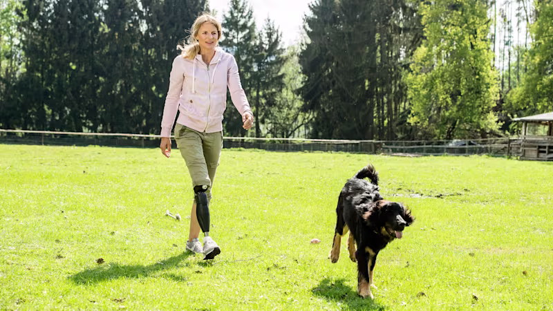 Auf dem Bild sieht man eine Frau, die auf einer Wiese mit ihrem Hund spazieren geht. Sie trägt eine Beinprothese, das Genium X3.