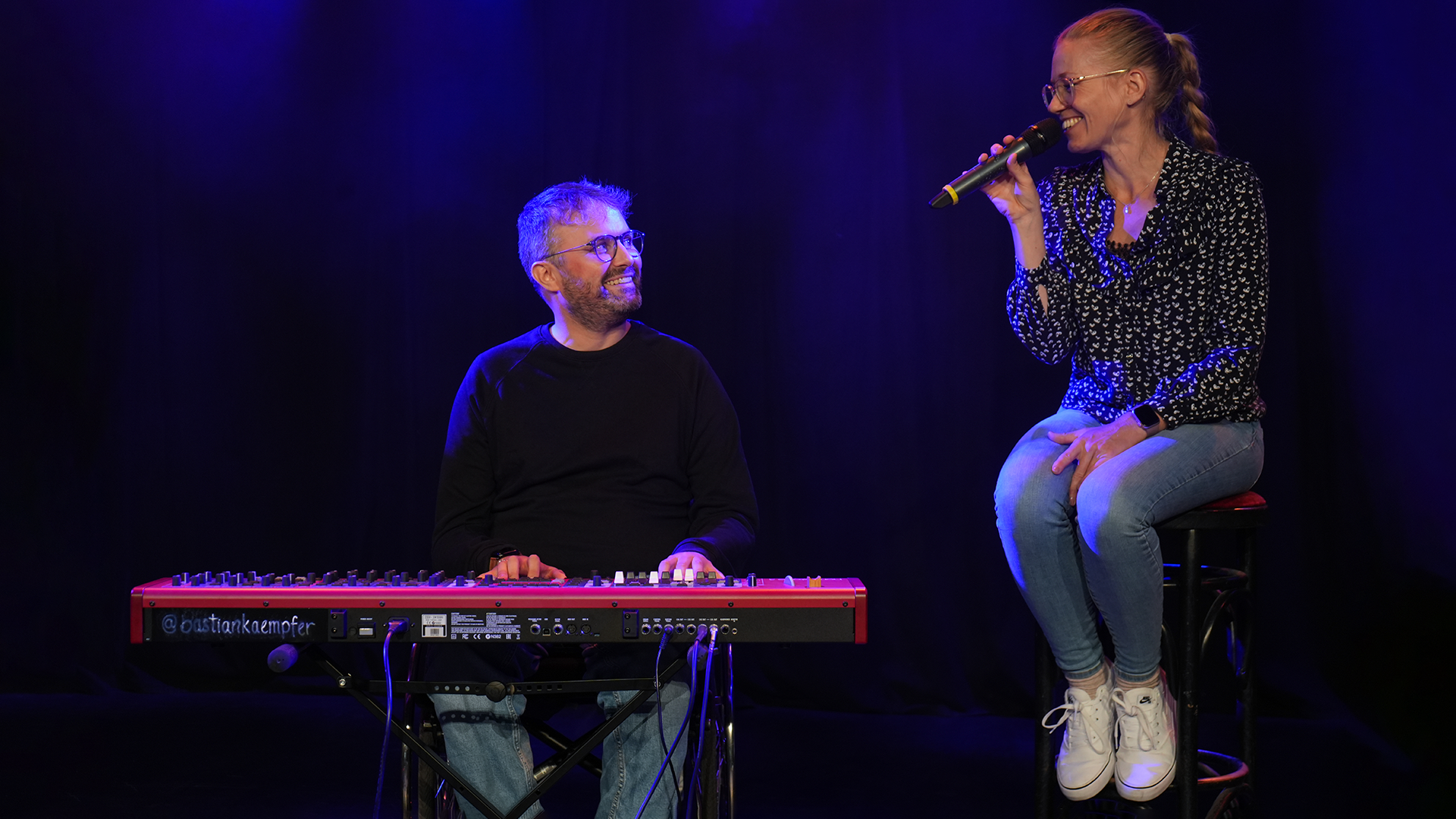 Eine männliche gelesene Person sitzt im Avantgarde Rollstuhl von Ottobock. Vor ihm steht ein Keyboard, auf dem seine Hände liegen. Er lächelt und guckt nach rechts zu einer weiblich gelesenen Person mit blonden Haaren. Sie sitzt auf einem Barhocker und hält ein Mikrofon in der Hand.