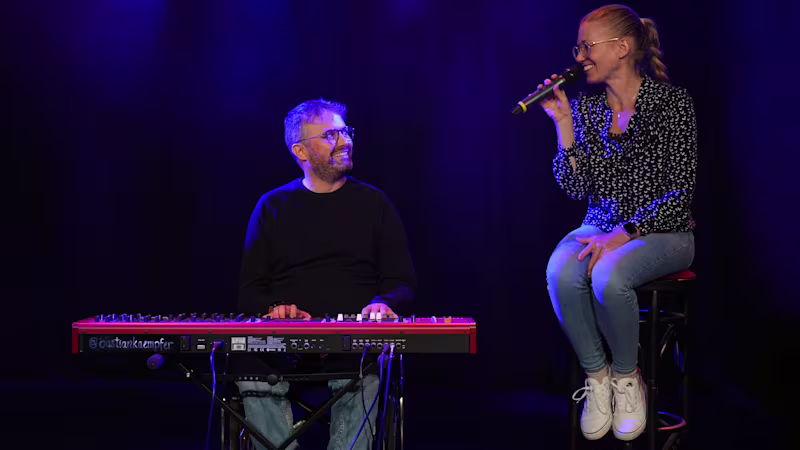 Eine männliche gelesene Person sitzt im Avantgarde Rollstuhl von Ottobock. Vor ihm steht ein Keyboard, auf dem seine Hände liegen. Er lächelt und guckt nach rechts zu einer weiblich gelesenen Person mit blonden Haaren. Sie sitzt auf einem Barhocker und hält ein Mikrofon in der Hand.