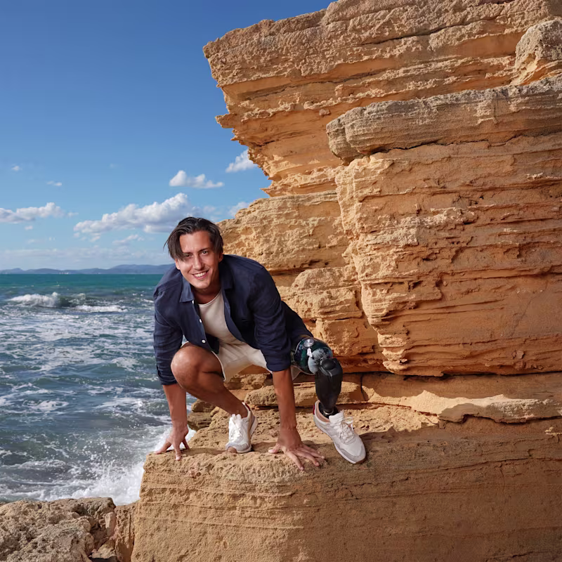 Alessandro Ossola with his Evanto prosthetic foot from Ottobock squatting down on a rock in front of the sea.