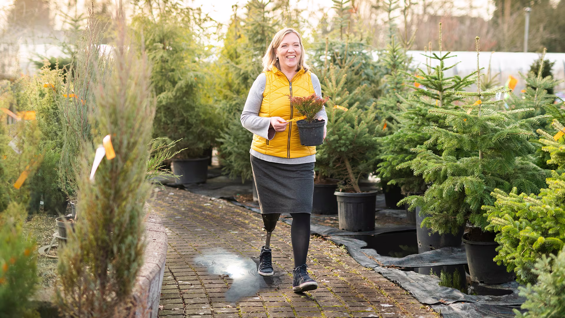 An amputee joyfully carries a potted plant through an outdoor garden. She is wearing the Ottobock Trias prosthetic foot.