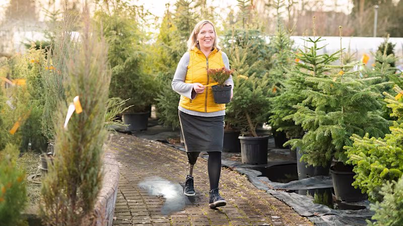An amputee joyfully carries a potted plant through an outdoor garden. She is wearing the Ottobock Trias prosthetic foot.