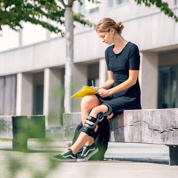 Eine weibliche Person sitzt auf einer Bank und trägt dabei die Genu Arexa.