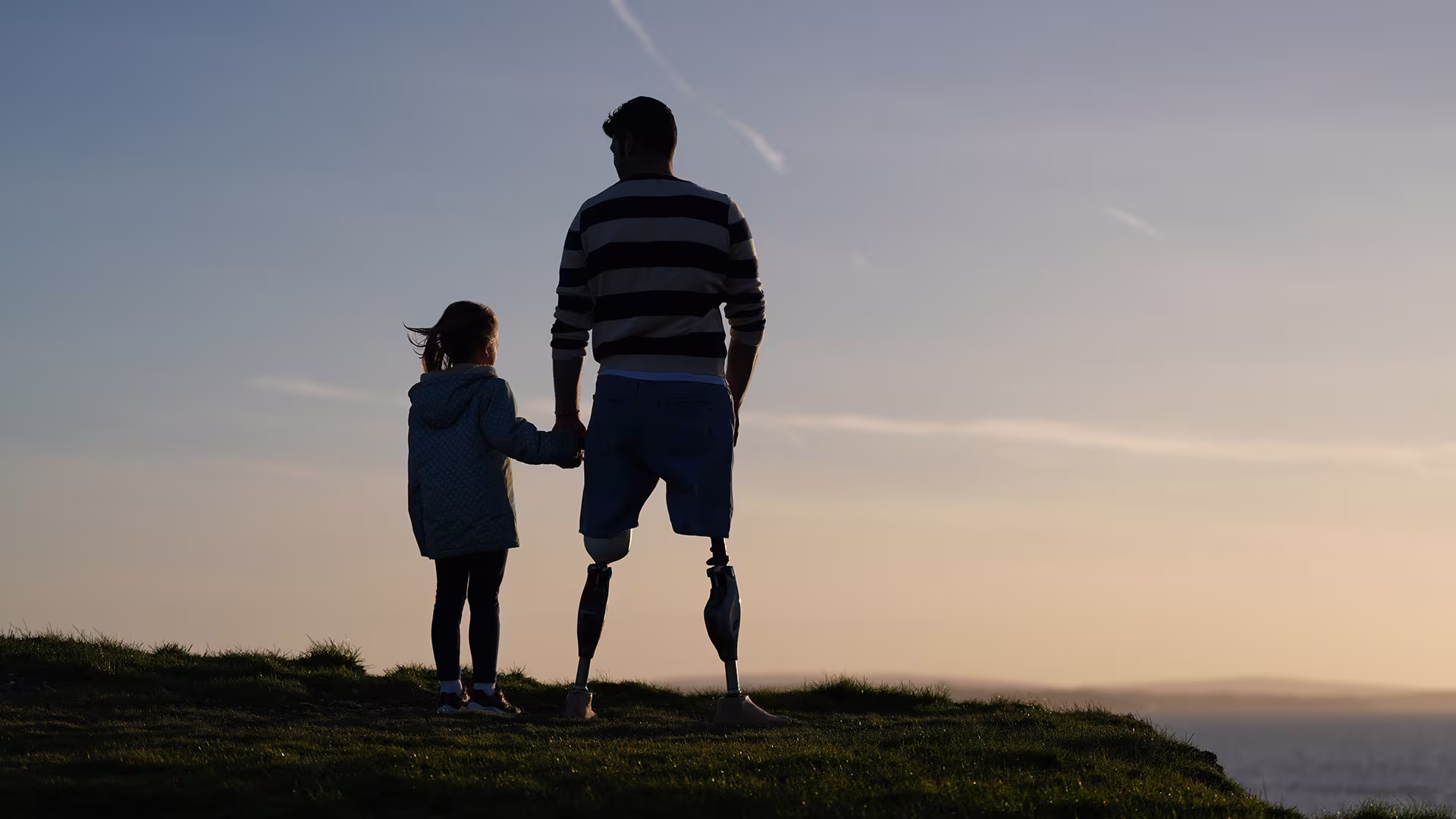 Bilateral leg amputee with Ottobock prosthetic legs stands at the coast holding hands with his daughter and his back turned to the camera, looking towards the sunset