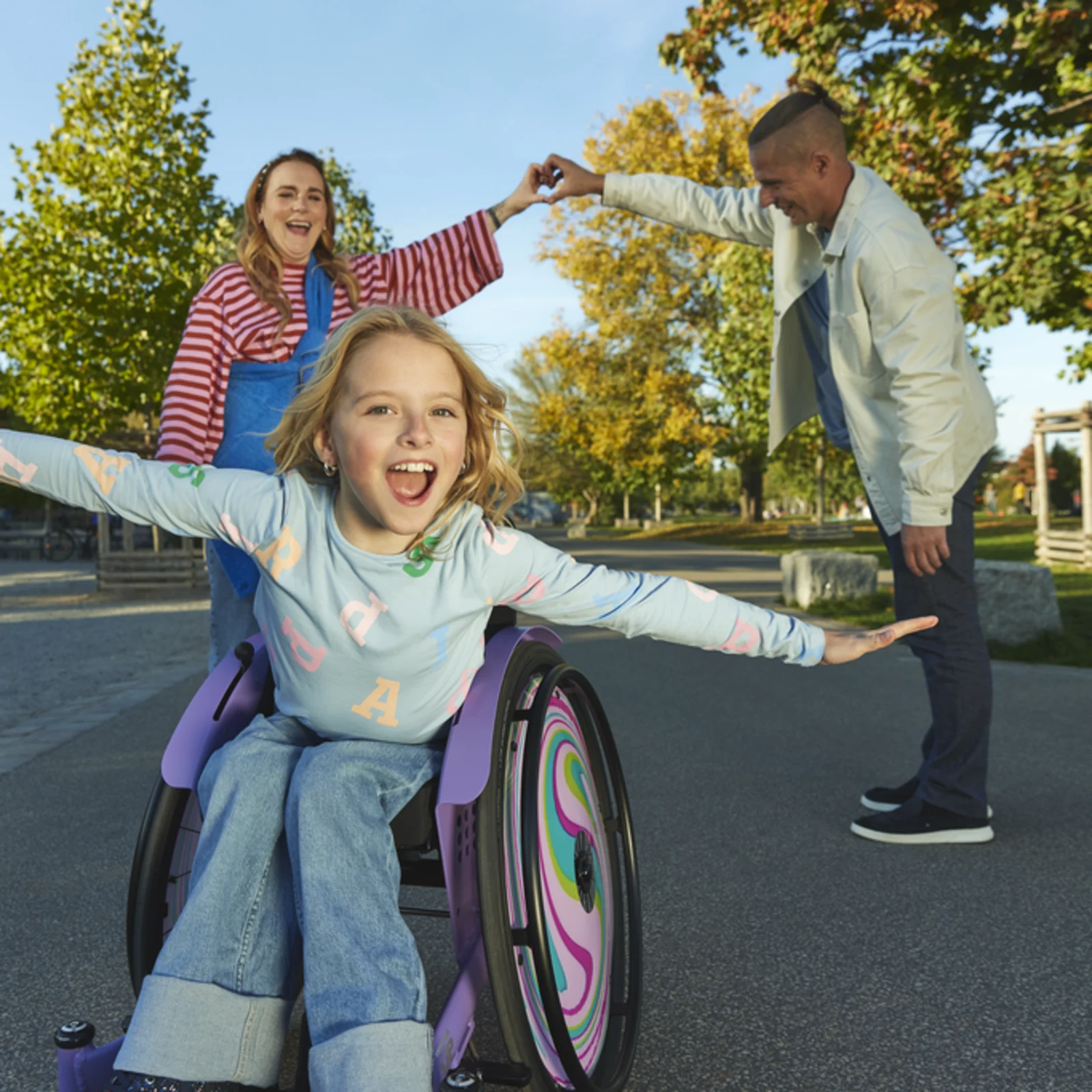 A young girl, Nomine, leans forward in her Kiddo wheelchair with her arms reaching out to the side, smiling as her parents’s stand behind her, hands joined to create a heart shape.