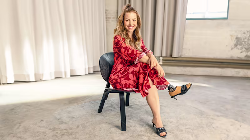 Eine junge Frau sitzt in einem hellen Raum auf einem Stuhl und lächelt in die Kamera. Sie hat die Beine überschlagen und trägt am rechten Fuß den Prothesenfuß Taleo Adjust von Ottobock.