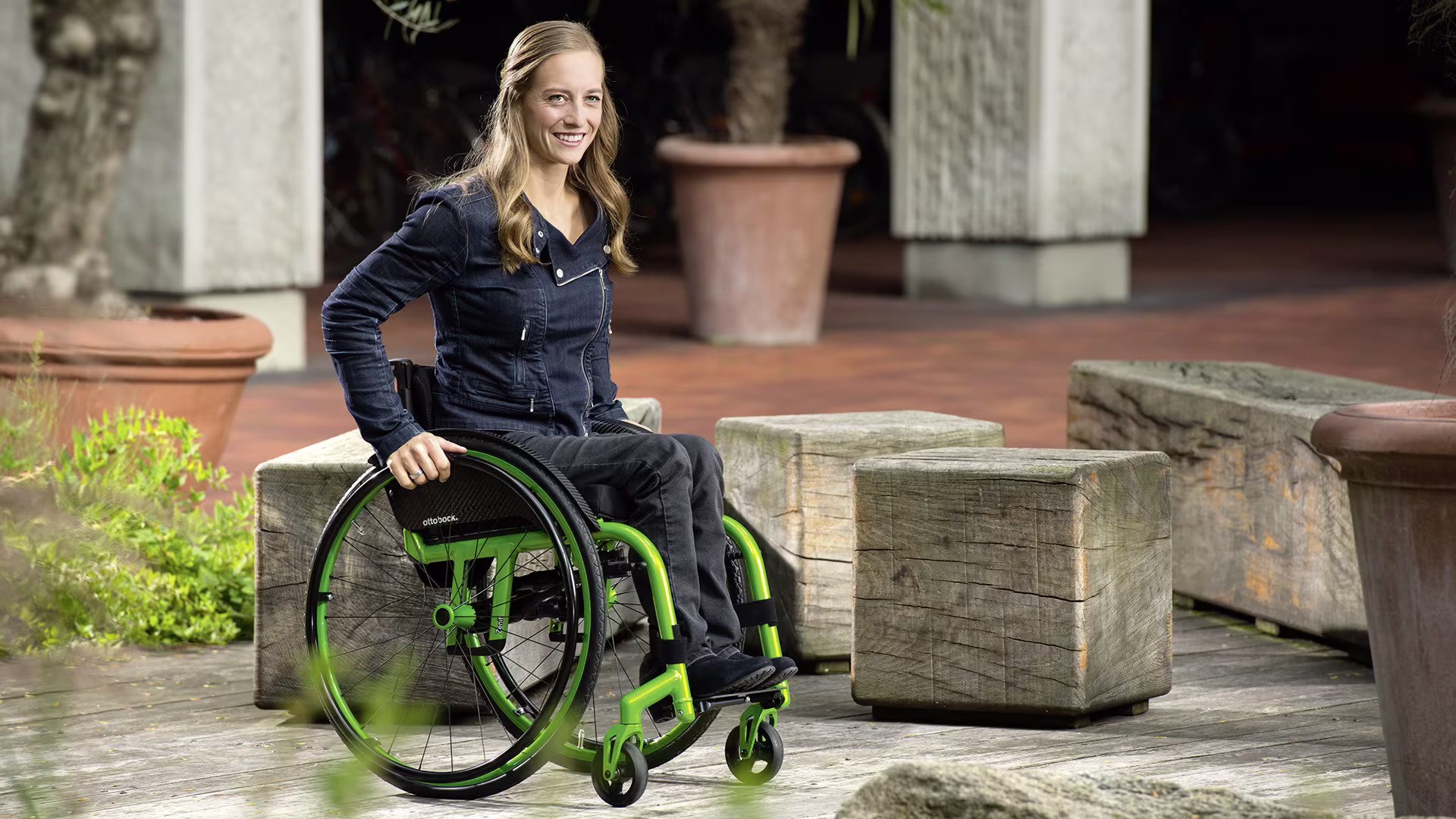 A lady in her Zenit CLT lightweight manual wheelchair from Ottobock is pushing in front of wooden benches.