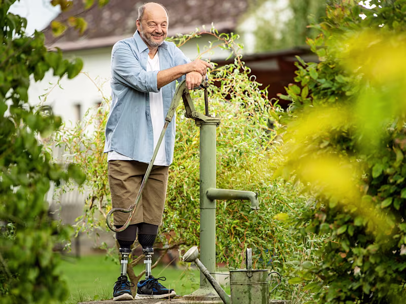 Person using prosthetic legs operates a green hand water pump in a lush garden, surrounded by plants and a watering can, with a house blurred in the background.