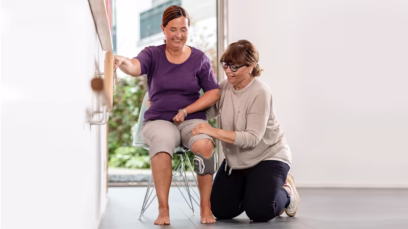 A woman sitting on a chair wearing a L300 Go Functional Electrical Stimulation device being helped by a physiotherapist.