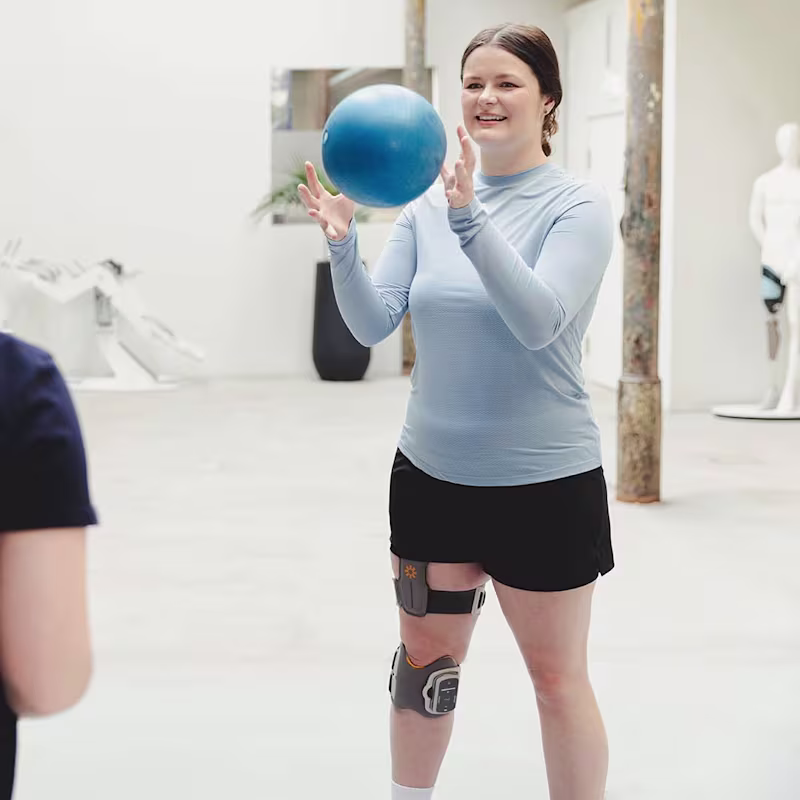 A woman read as a woman stands on a balance board. She is wearing Ottobock’s L300 Go on her right leg. She tosses a blue ball back and forth with another female read as a woman.