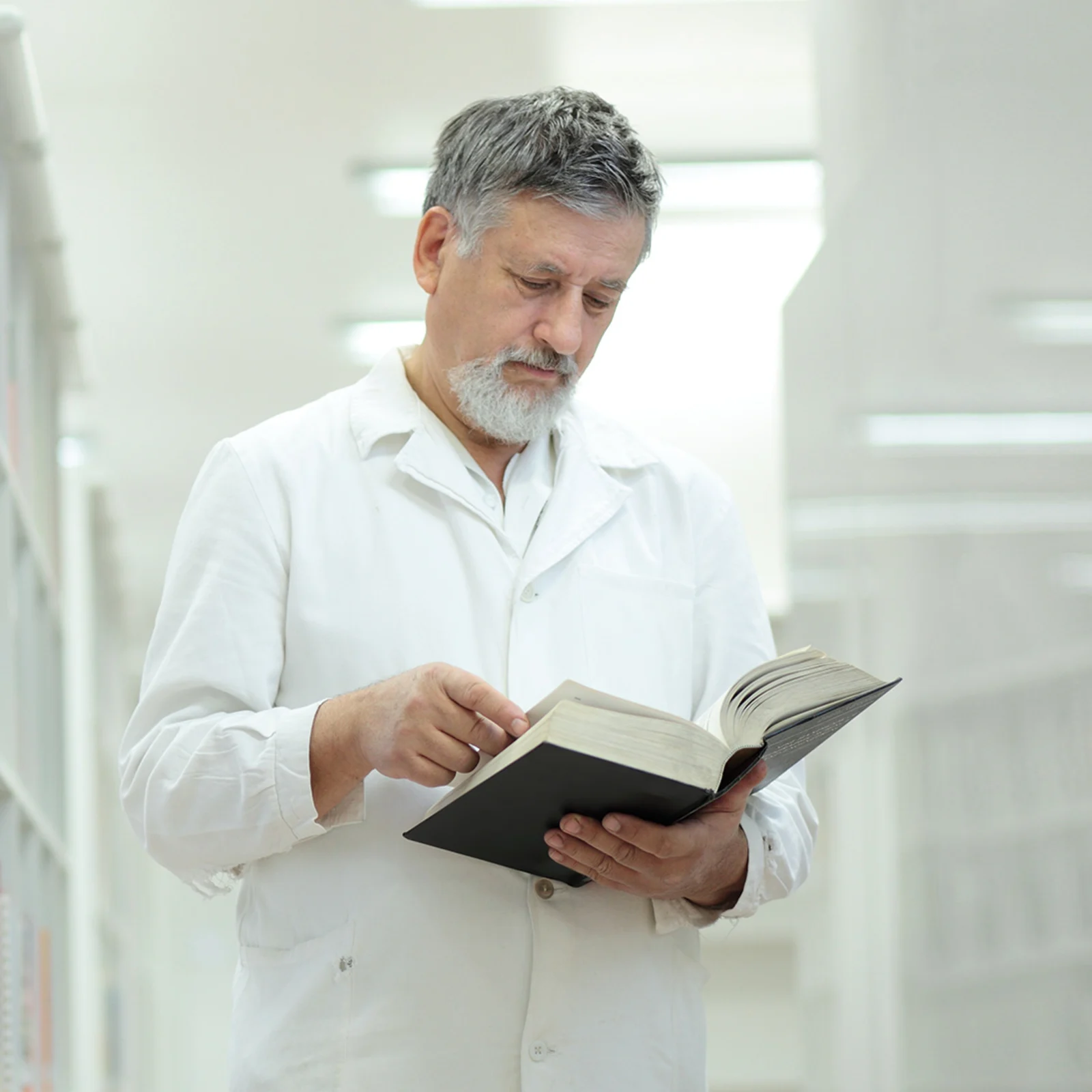 Eine männlich gelesene Person im Arztkittel liest ein Buch in der Bibliothek. 