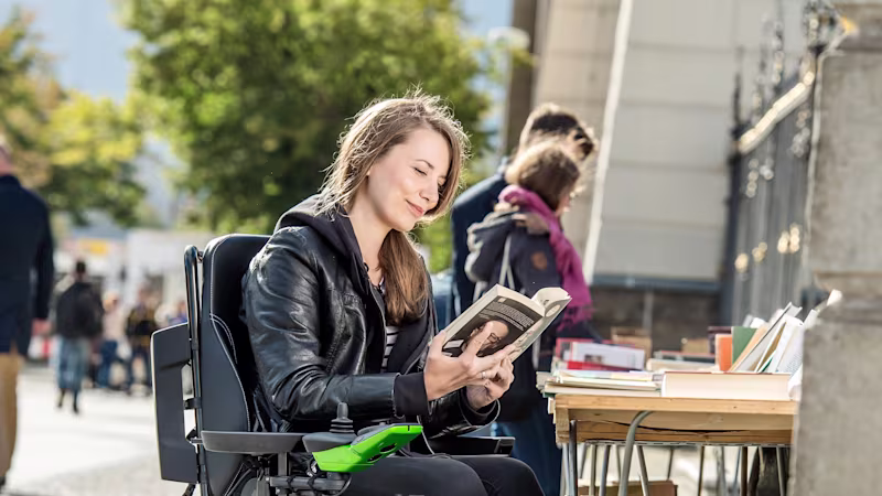 Tekerlekli sandalye kullanıcısı Kristina, Ottobock Juvo B4'te sokaktaki kullanılmış kitap standında oturuyor ve kitap okuyor.