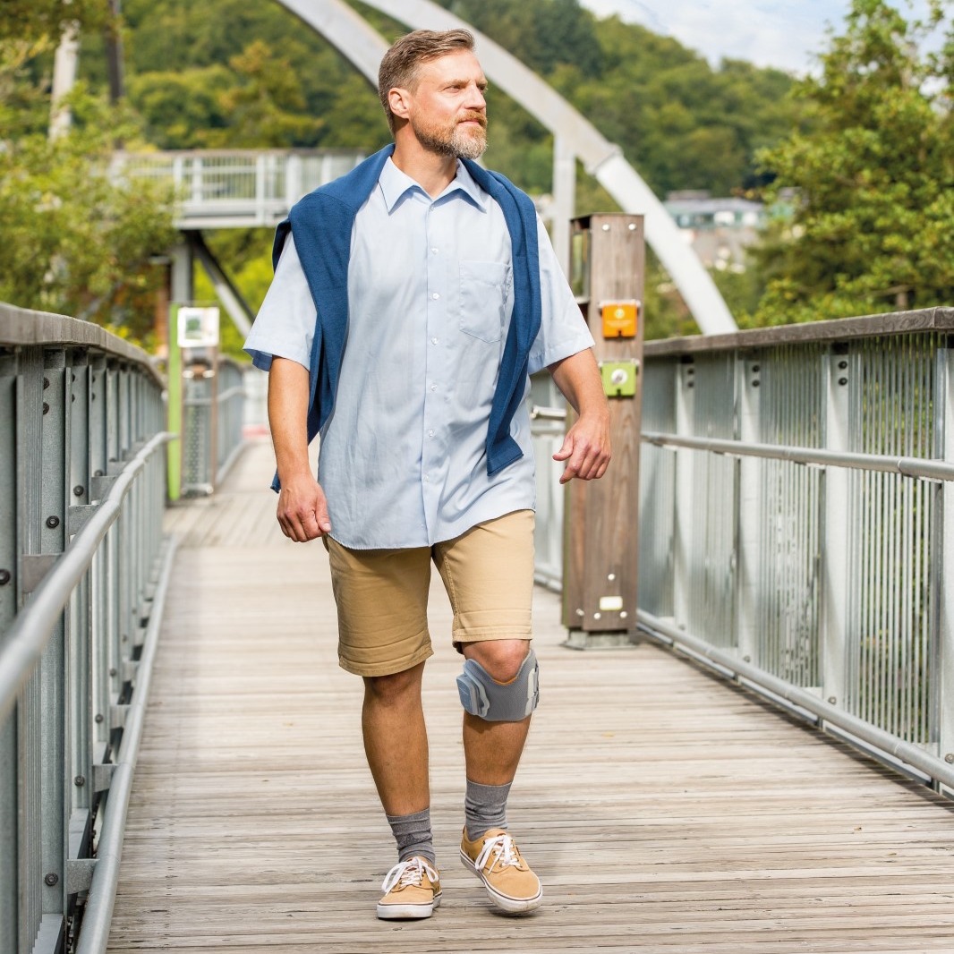 Dirk, a man recovering from a stroke, walks outdoors across a bridge wearing an FES device on his leg. The functional electrical stimulation system supports his mobility and helps improve natural walking movement during rehabilitation.
