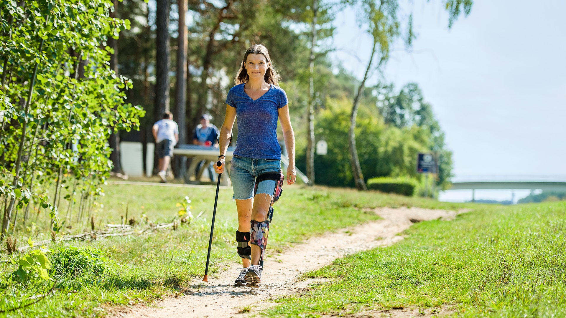 A woman wearing an E-MAG Active & Nexgear Tango ankle joint walking with a crutch in a park. 