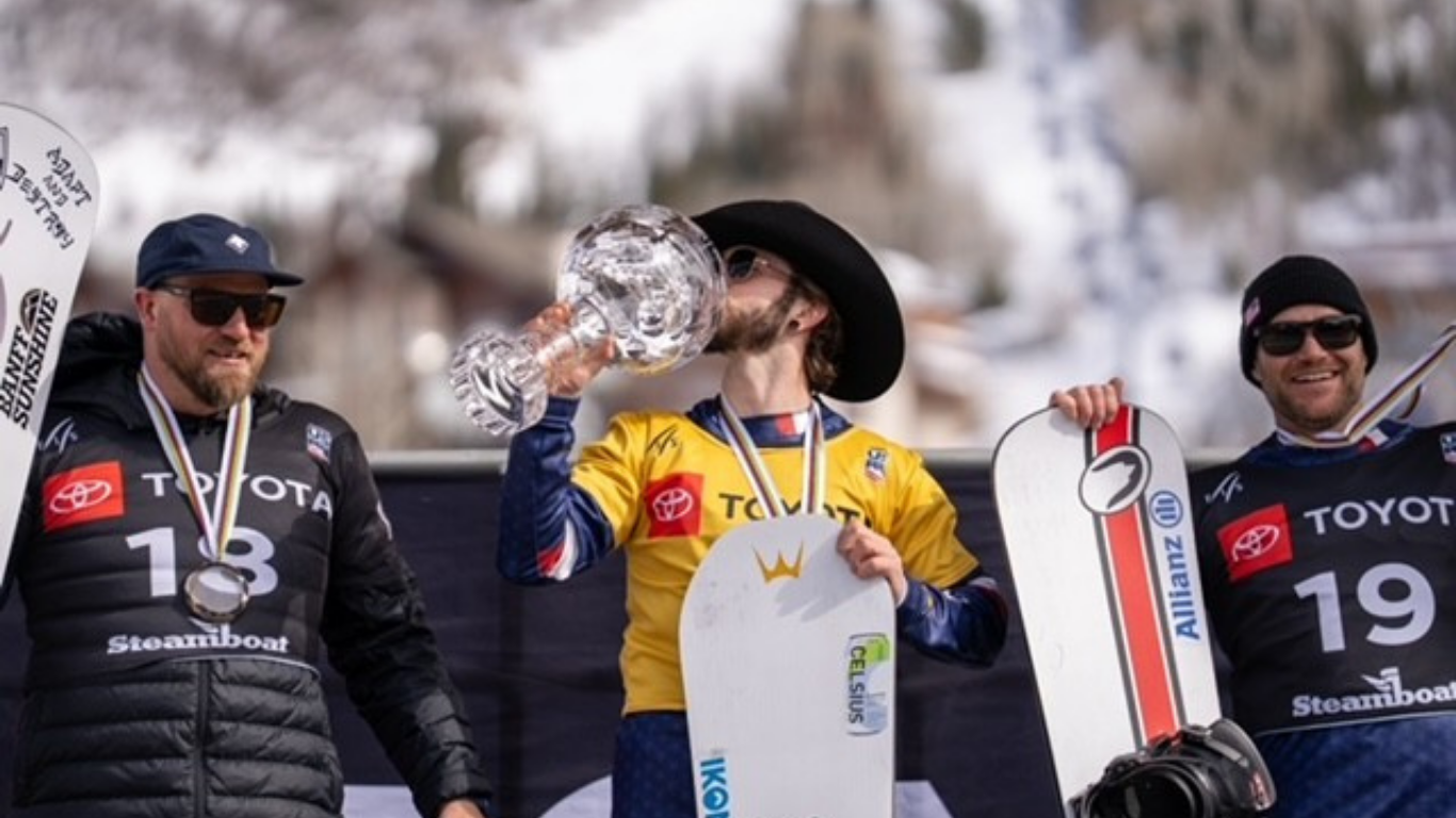 Three men stand on the podium with medals around their necks after a snowboarding event. The first place winner kisses the trophy while wearing a black cowboy hat