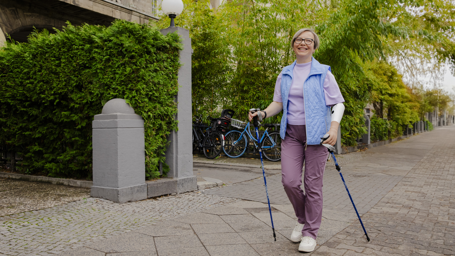 Auf dem Bild sieht man Bozena. Sie geht lächelnd draußen mit Nordic-Walking Stöcken spazieren. Sie trägt eine myoelektrische Armprothese mit der Speedhand Handprothese.