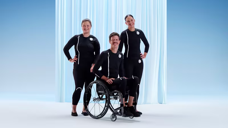 Auf dem Bild sieht man rechts und links zwei weiblich gelesene Frauen die den exopulse suit tragen. In der Mitte sitzt ein junger Mann im Rollstuhl und trägt ebenfalls den exopulse suit. Alle drei lächeln in die Kamera.