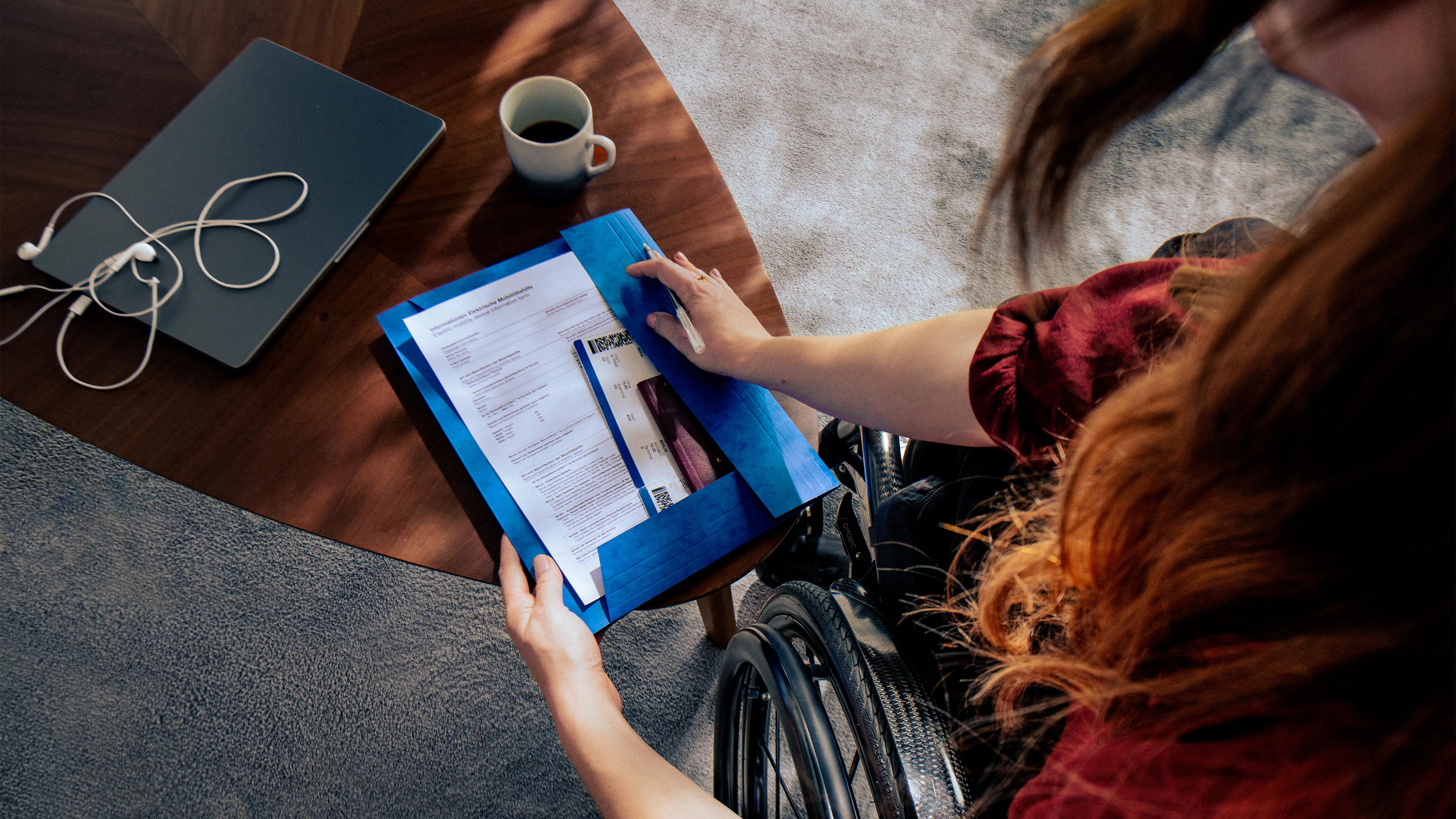 Person using a wheelchair reviewing travel documents at a desk with a laptop and coffee, illustrating the extra bureaucracy and planning challenges shown in Ottobock’s Invisible Class campaign.