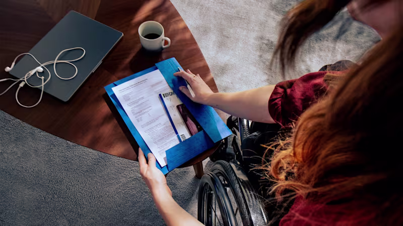 Person using a wheelchair reviewing travel documents at a desk with a laptop and coffee, illustrating the extra bureaucracy and planning challenges shown in Ottobock’s Invisible Class campaign.