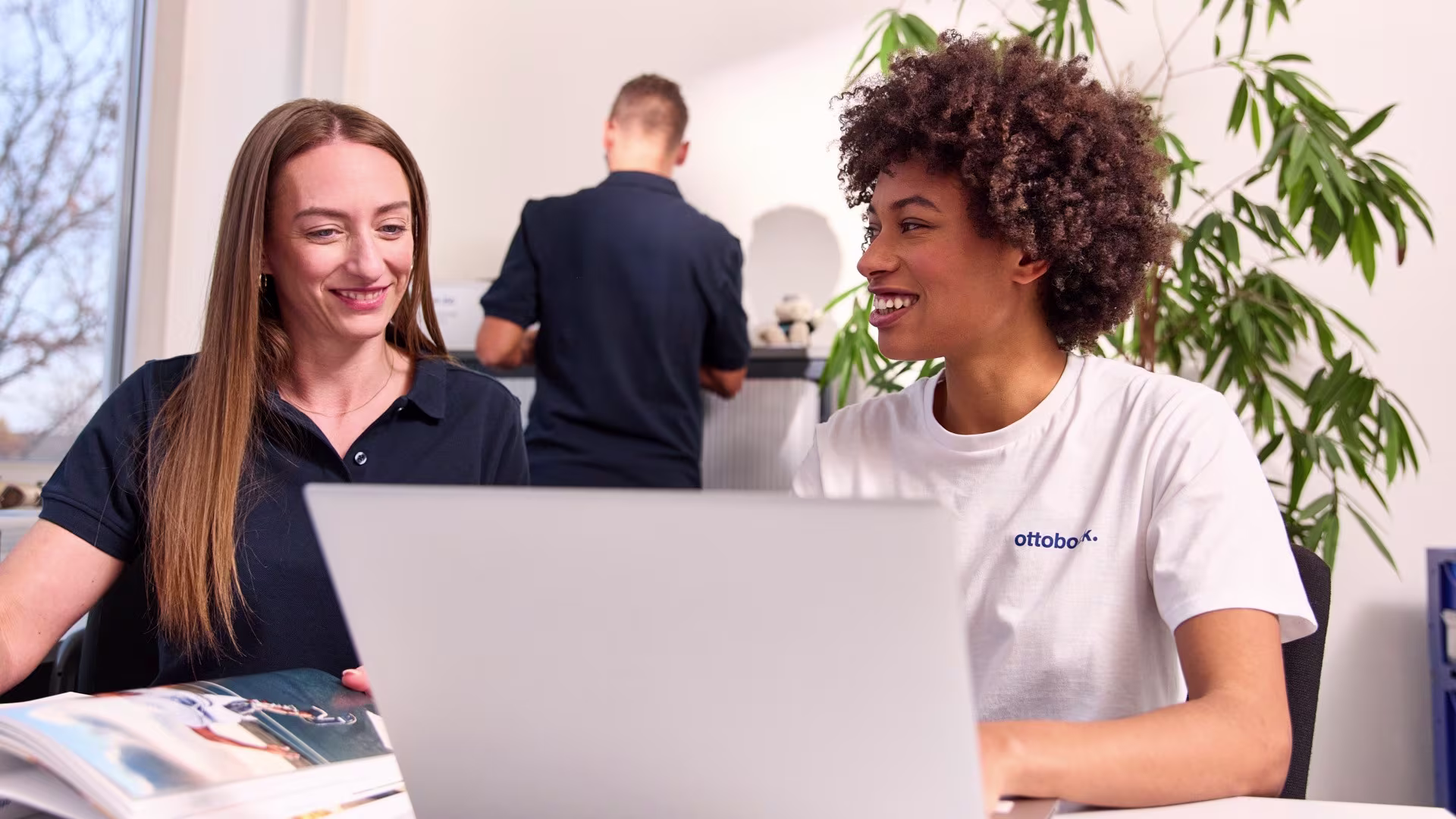 Two Ottobock employees sitting together at a laptop in an office discussing documents, with a colleague working at a printer in the background.