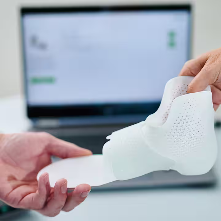 Hands holding 3D-printed orthotic device with laptop in background at Ottobock workspace.
