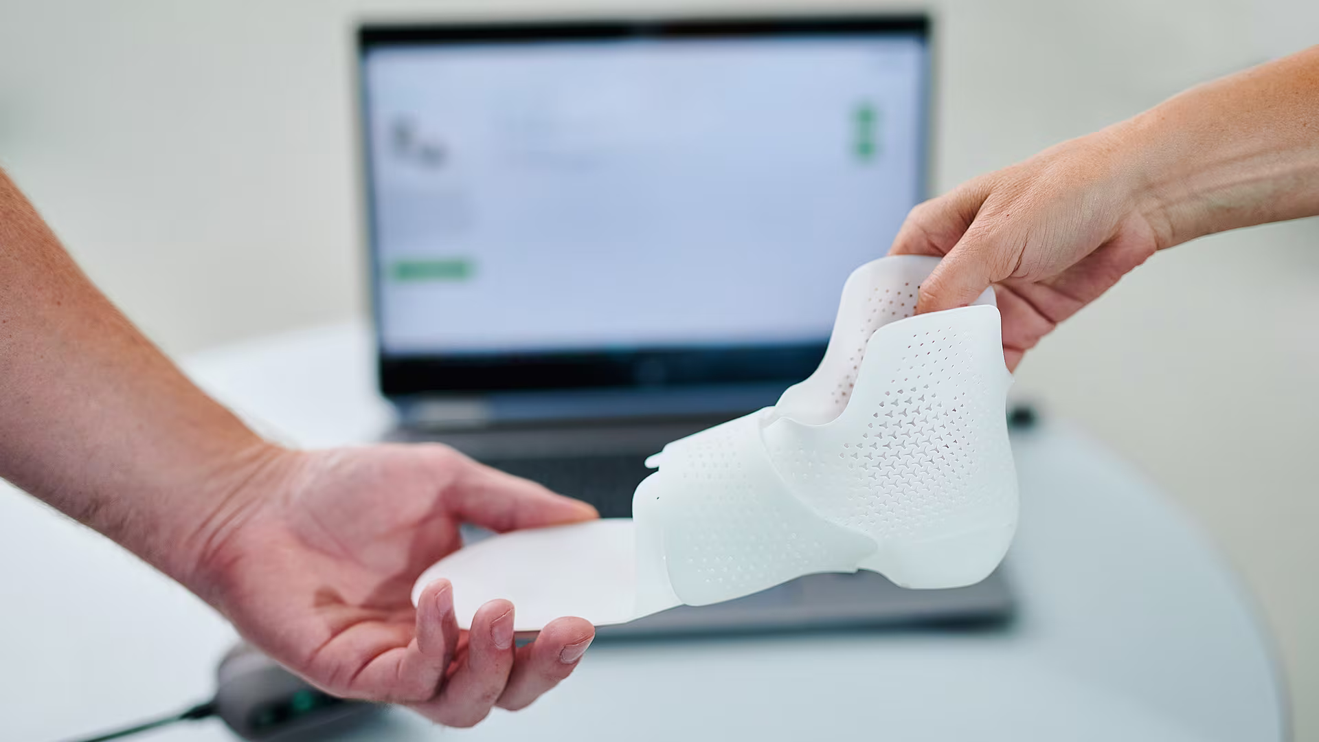 Mani che tengono dispositivo di ortotica stampato in 3D con computer portatile sullo sfondo nell’area di lavoro Ottobock.