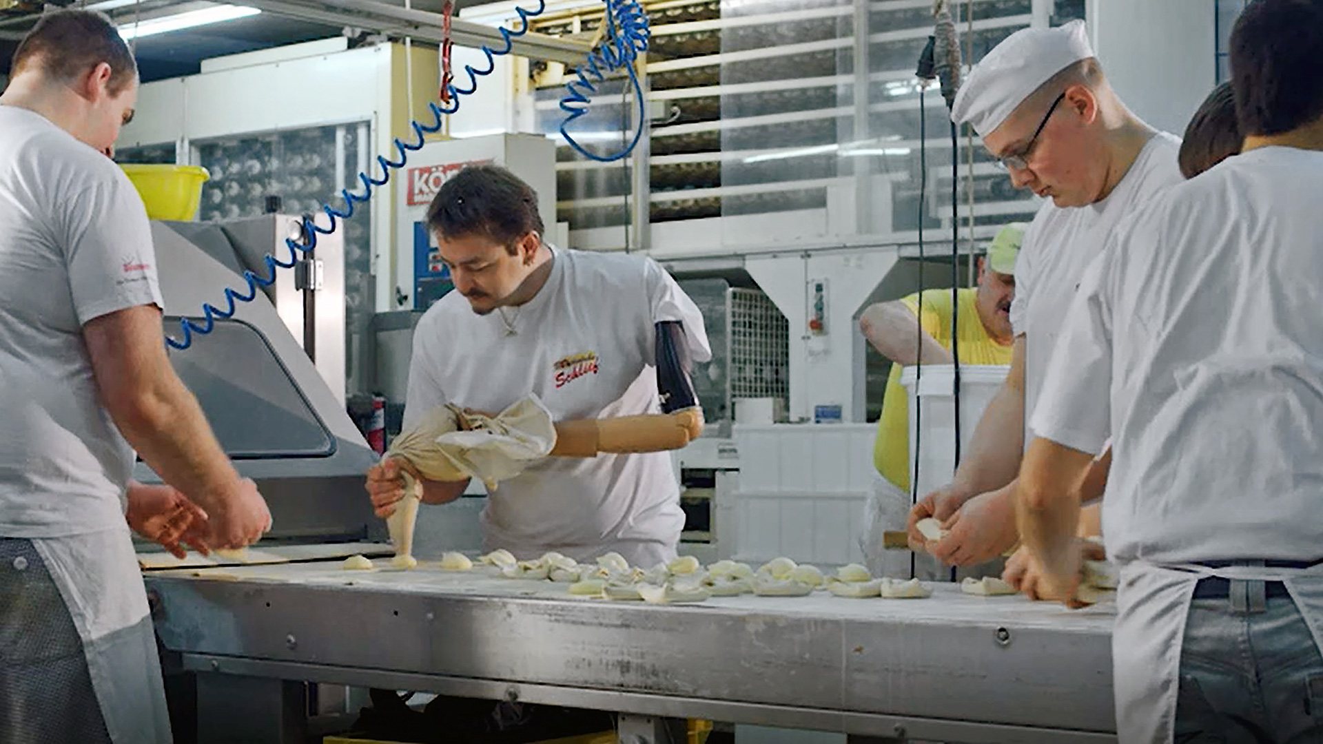 Auf dem Bild sieht man vier Männer die an einem Fließband in einem Bäckereibetrieb stehen. Der Mann in der Mitte trägt eine Armprothese und spritzt gerade mit einem Spritzbeutel eine Masse auf das Fließband. 
