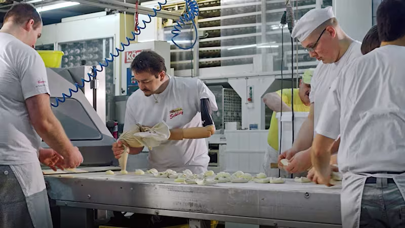 Auf dem Bild sieht man vier Männer die an einem Fließband in einem Bäckereibetrieb stehen. Der Mann in der Mitte trägt eine Armprothese und spritzt gerade mit einem Spritzbeutel eine Masse auf das Fließband.
