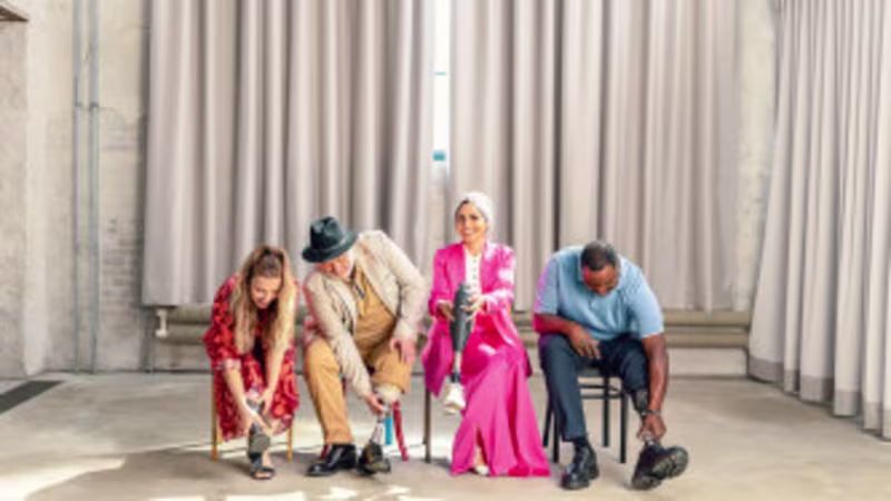 Vier Ottobock Fußprothesenträger sitzen in einer Reihe in einem Raum: zwei Frauen und zwei Männer, alle probieren unterschiedliche Schuhe aus und lachen.