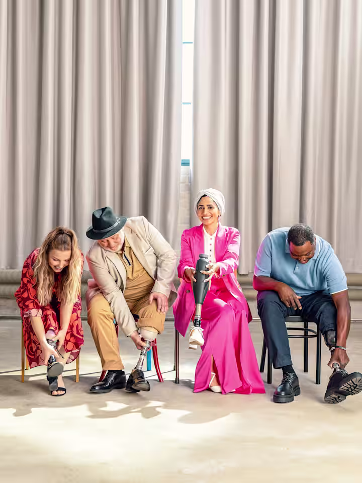Four Ottobock prosthetic foot users sitting in a row in a clean room: two women and two men, all trying different shoes and laughing.