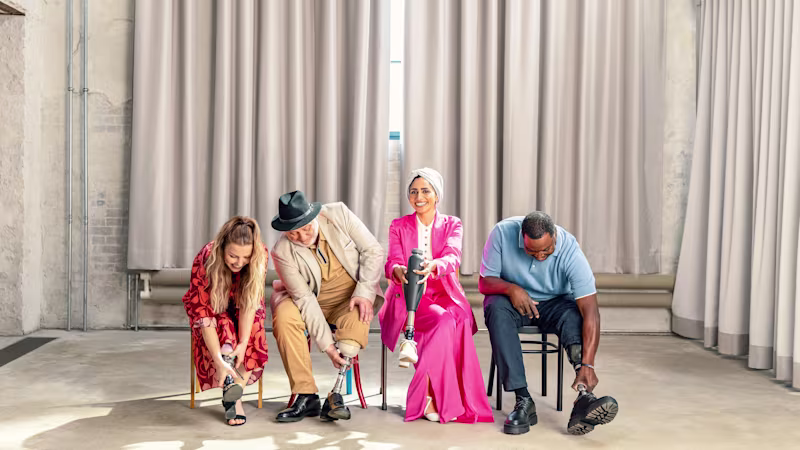 Four Ottobock prosthetic foot users sitting in a row in a clean room: two women and two men, all trying different shoes and laughing.