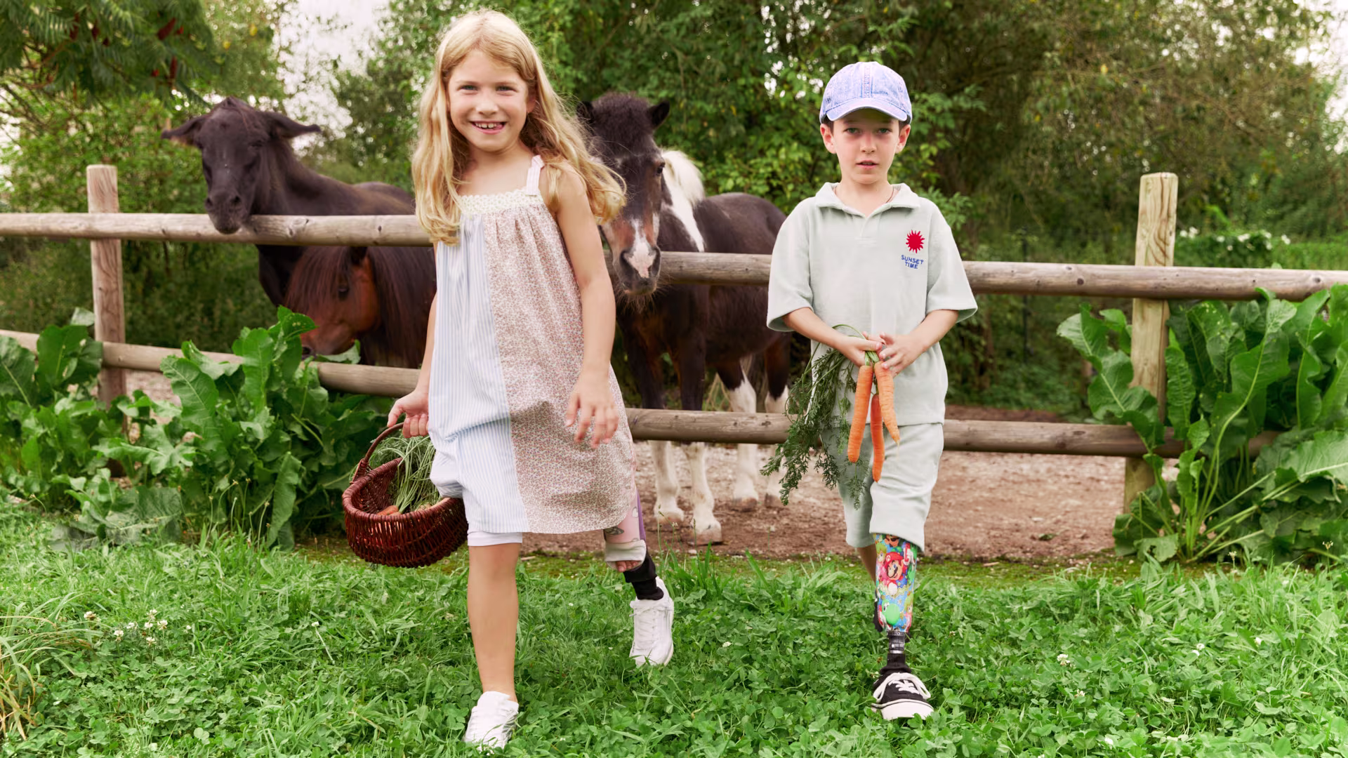 Two children with Ottobock leg prostheses in a stable carrying a basket of carrots with horses in the background.