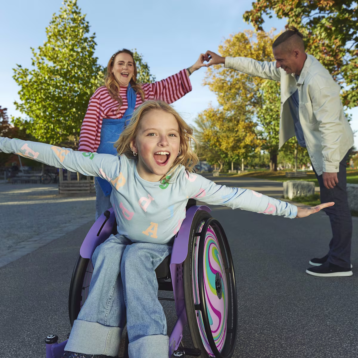 A young girl sitting in her kidevo wheelchair with her arms outstretched (as if she was flying) and her parents form a heart with their hands in the background.