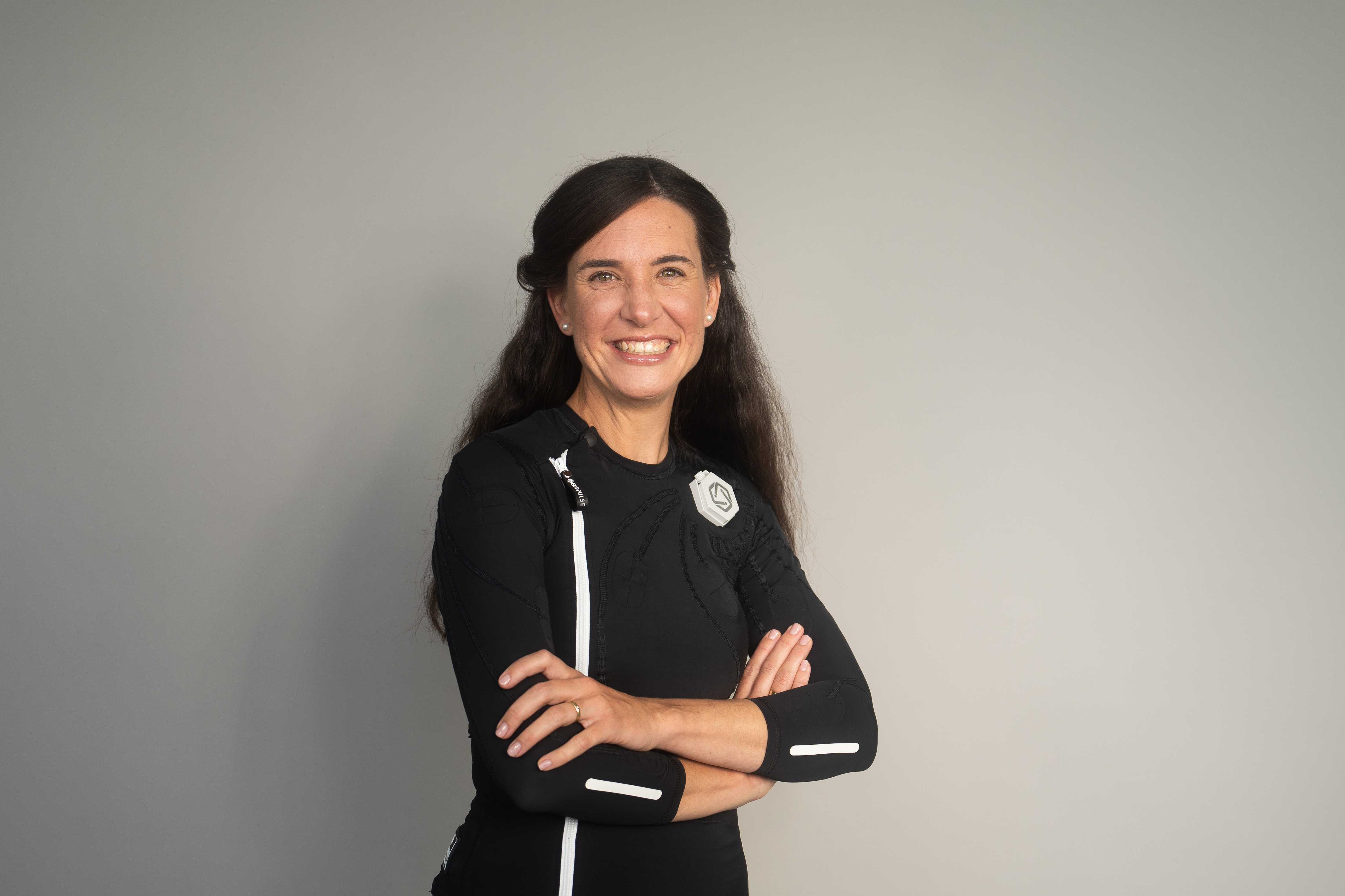 Eine dunkelhaarige Frau trägt den exopulse suit von Ottobock. Sie sitzt auf einem beigen Sofa und lächelt in die Kamera.