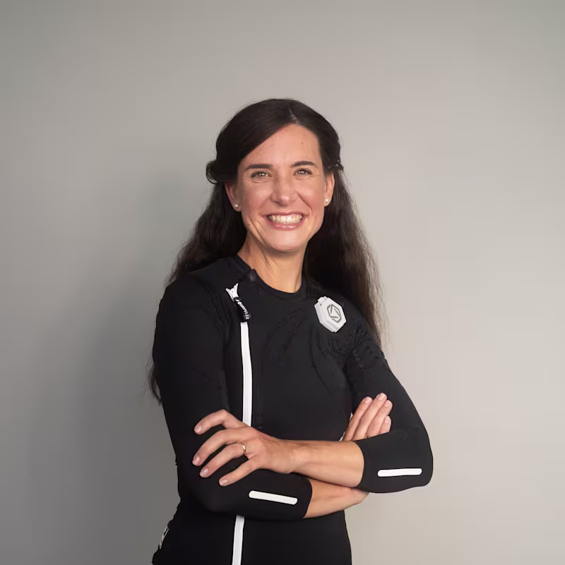 Eine dunkelhaarige Frau trägt den exopulse suit von Ottobock. Sie sitzt auf einem beigen Sofa und lächelt in die Kamera.