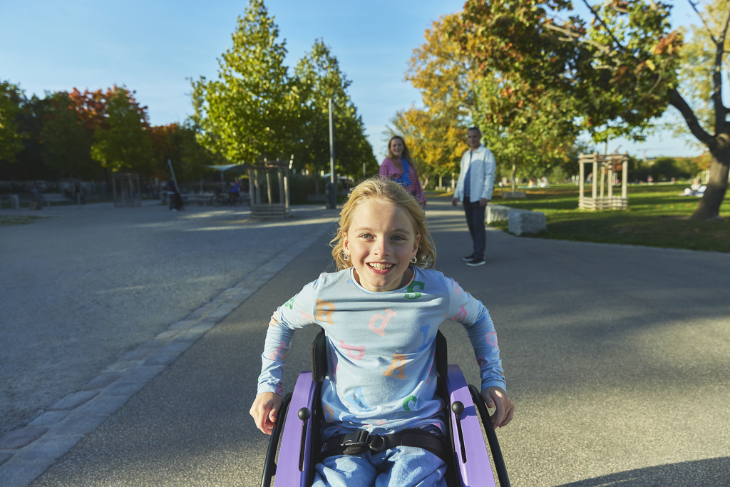 In the foreground, a young girl, Nomine, is outside, smiling widely and seated in her kidevo wheelchair as her parents look on in the background.