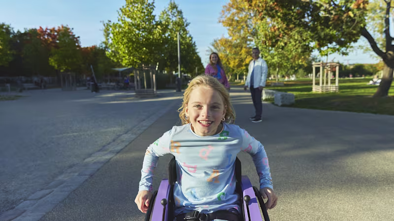 Ön planda, Nomine adında genç bir kız dışarıda, kidevo tekerlekli sandalyesinde oturmuş, gülümsüyor ve arka planda ebeveynleri ona bakıyor.
