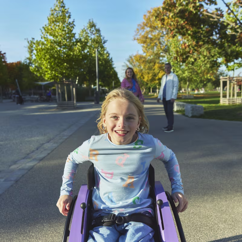 Ön planda, Nomine adında genç bir kız dışarıda, kidevo tekerlekli sandalyesinde oturmuş, gülümsüyor ve arka planda ebeveynleri ona bakıyor.