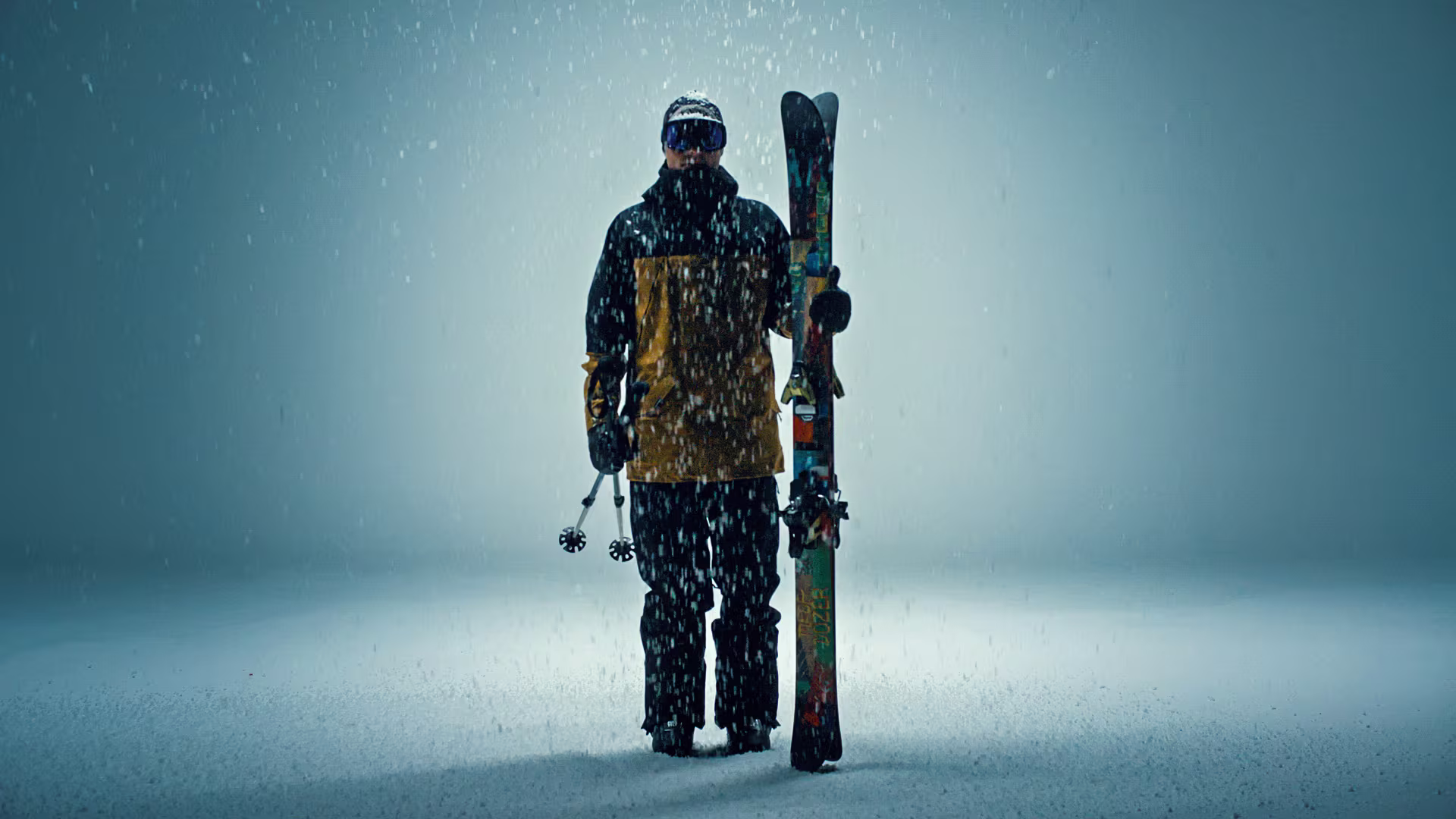 Sciatore in completo abbigliamento da sci che tiene gli sci nella mano sinistra e in piedi di fronte a uno sfondo scuro.