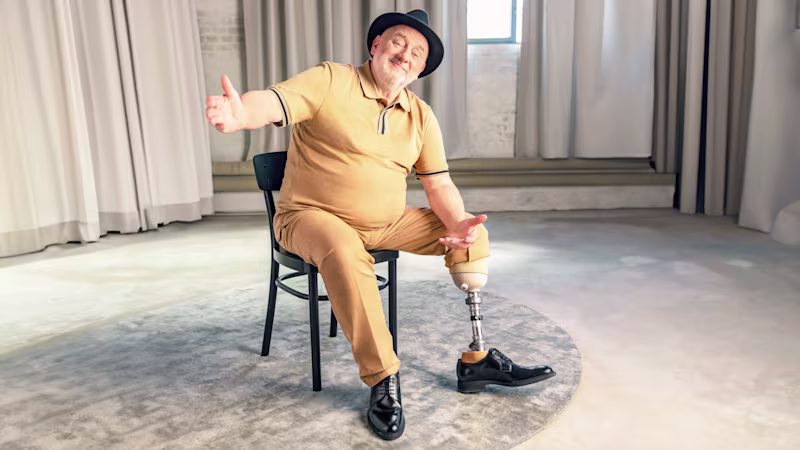Ein älterer Mann sitzt auf einem Stuhl, lächelt in die Kamera und streckt dabei einladend seine Hand aus. Er trägt den Quickchange Adapter und den Trias Prothesenfuß von Ottobock.