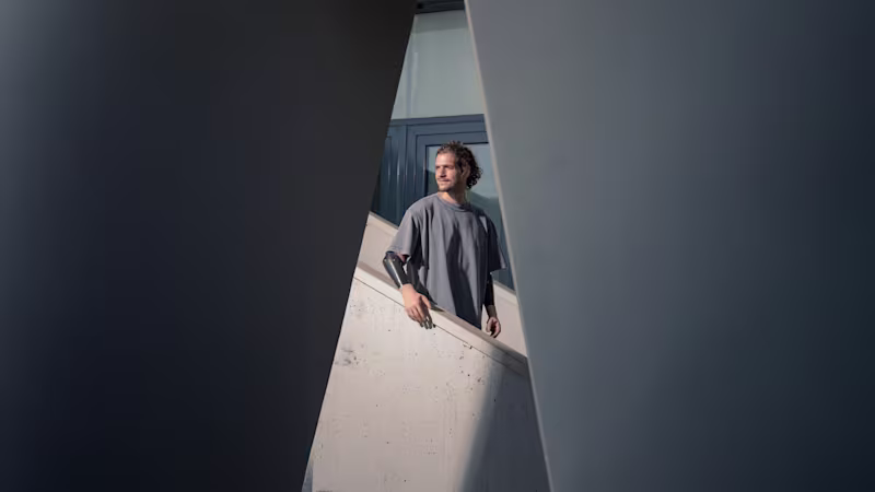 Homem com braços protéticos em pé entre estruturas arquitetónicas, simbolizando resiliência e independência na campanha Invisible Class da Ottobock sobre ultrapassar barreiras de viagem para pessoas com deficiência.