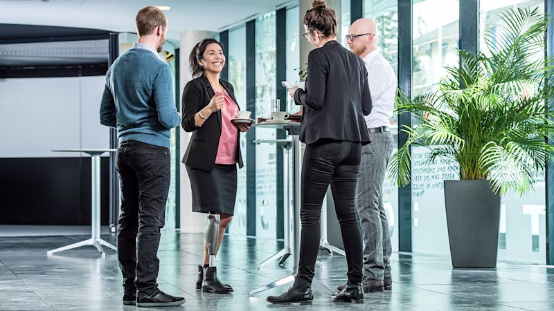 Vier Personen stehen an einem Stehtisch und reden miteinander. Im Fokus steht eine weiblich gelesene Person, die ein C-Leg trägt.