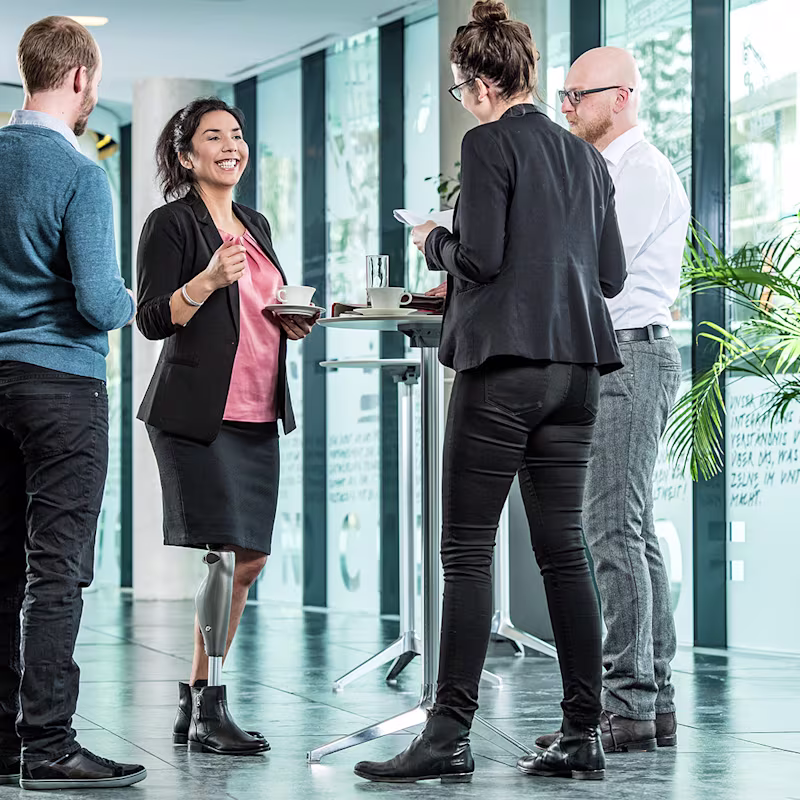 Vier Personen stehen an einem Stehtisch und reden miteinander. Im Fokus steht eine weiblich gelesene Person, die ein C-Leg trägt.